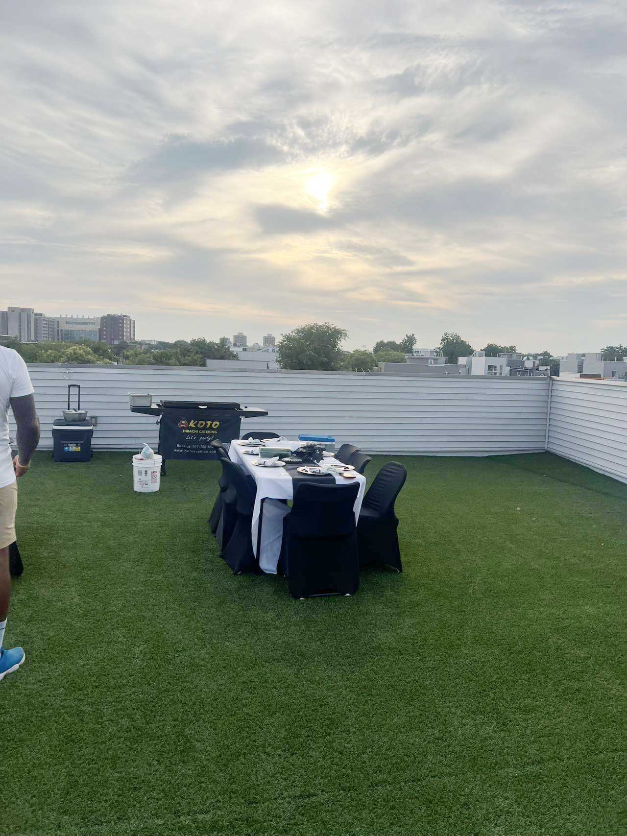 Rooftop catering event scene with artificial green turf, set dining table with white cloth and black chair covers. Koto hibachi catering grill station, white perimeter wall, distant city skyline under soft cloudy sunset sky.