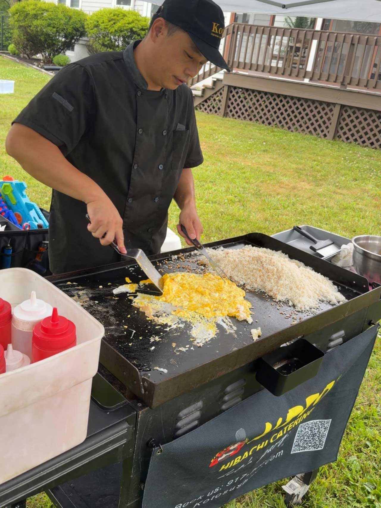Catering chef in black uniform and cap stir‑fries scrambled eggs and rice on outdoor hibachi grill. Squeeze sauce bottles sit nearby, branded catering banner, green grass lawn and residential backyard setting.