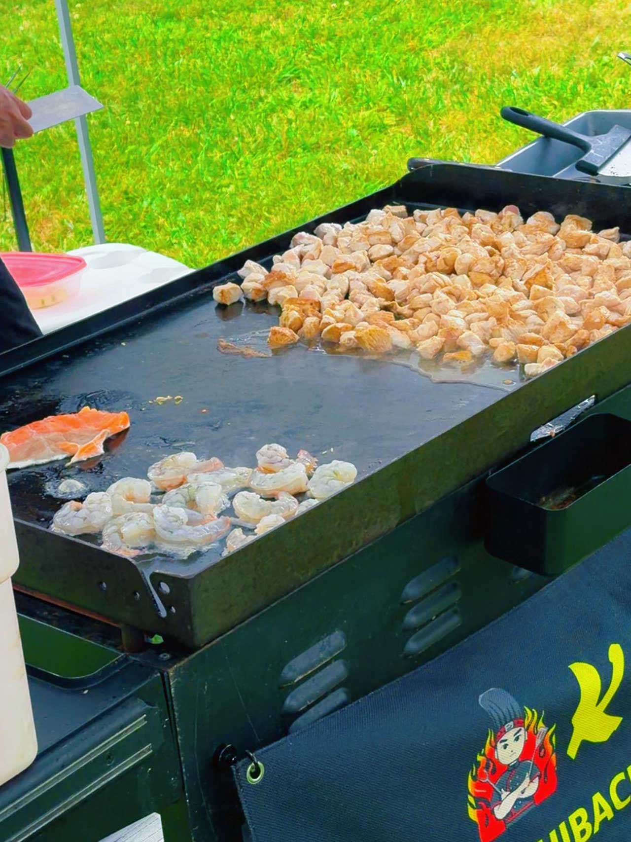 Outdoor hibachi flat‑top grill cooking raw shrimp and diced chicken pieces. Seafood and poultry spread across hot metal surface, branded catering banner visible, bright green grass lawn in the background.