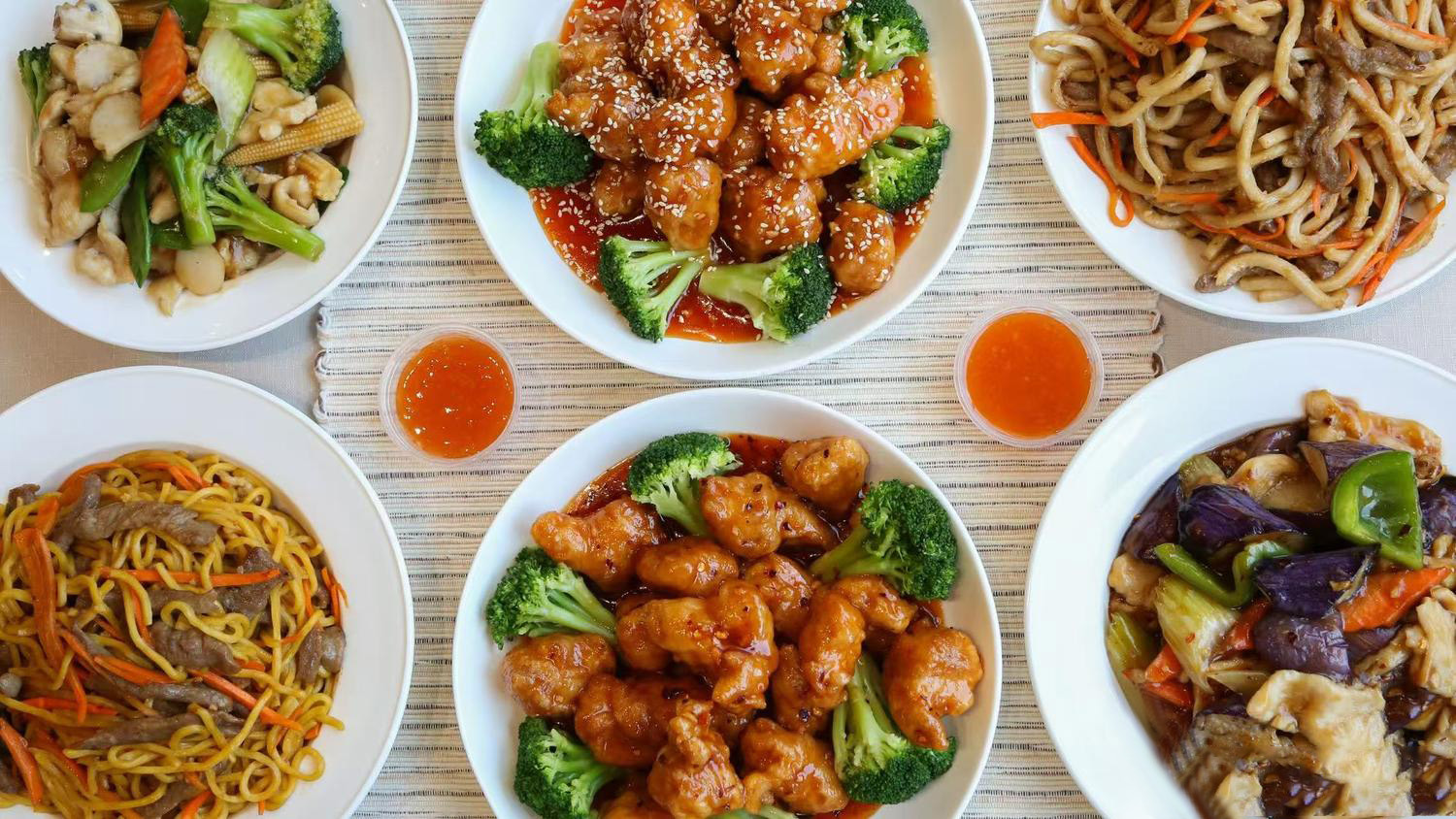 Top-down spread of Chinese dishes sesame chicken with broccoli, beef lo mein, vegetable stir-fry, and small sauce cups