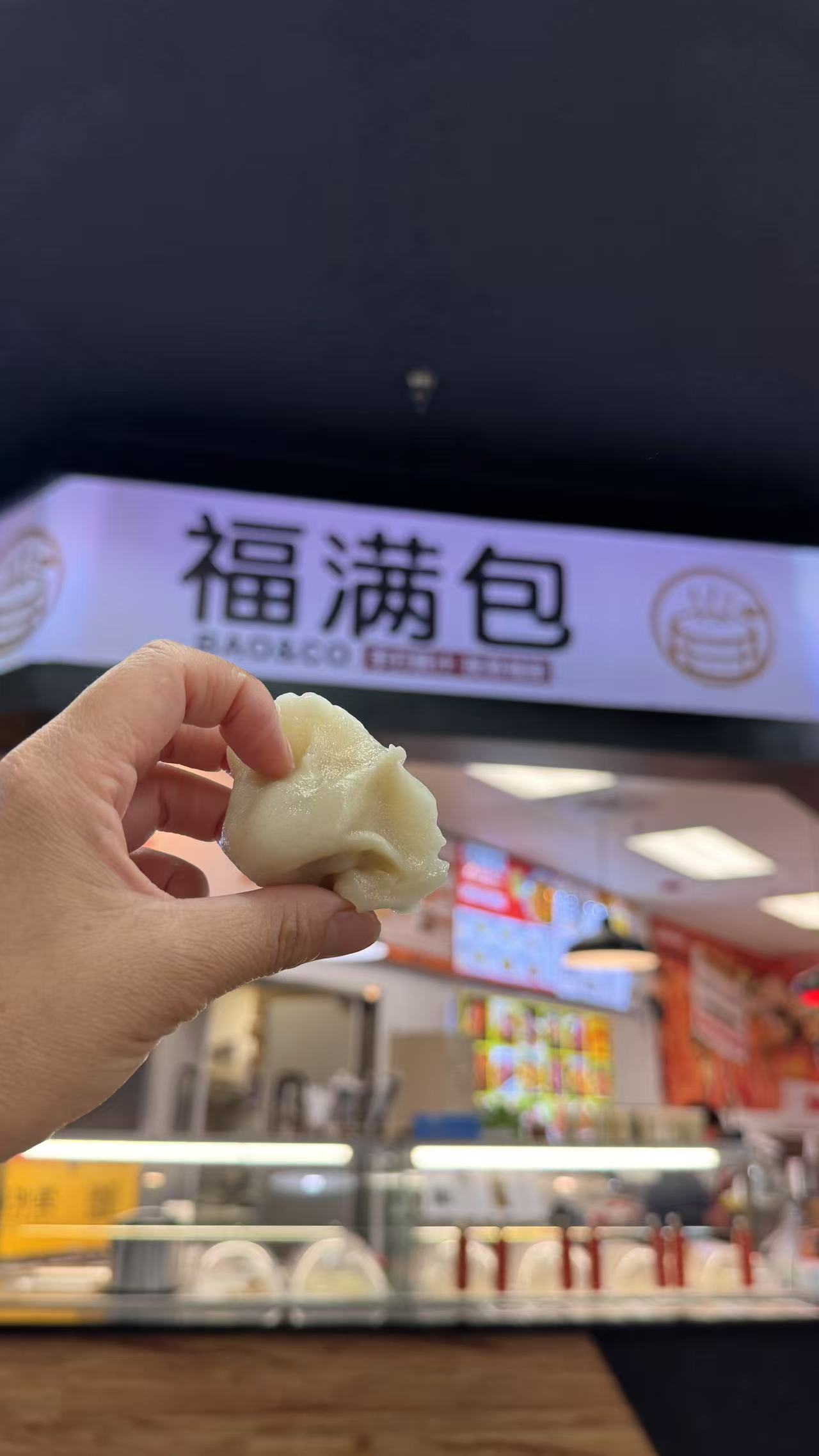 A hand holding a freshly made dumpling in front of the BAO CO restaurant sign, showcasing its soft and plump appearance