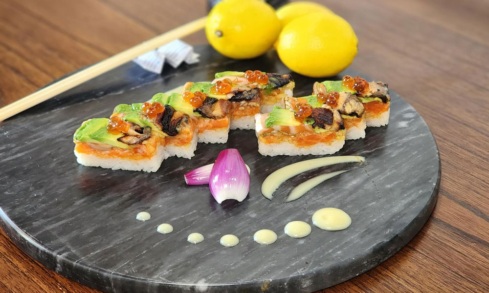 Sliced square sushi with salmon, avocado, eel and salmon roe, plated on dark marble stone board with onion garnish, sauce dots and chopsticks.
