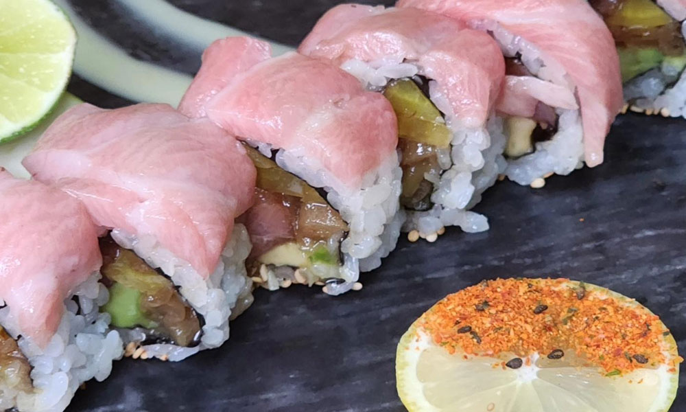 Sushi roll wrapped with fresh tuna slices, filled with avocado and pickled veggies, served on dark stone plate with spiced lime slice.