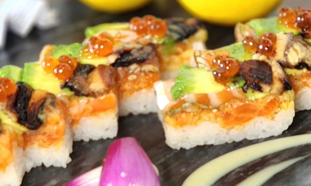Close view of square sushi pieces topped with salmon, avocado, grilled eel and orange salmon roe, decorated with purple onion and creamy sauce.