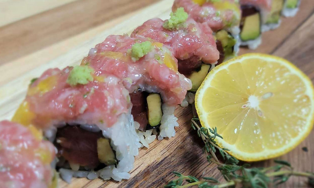 Sushi roll covered with chopped raw tuna, wasabi and mango, avocado filling, placed on wooden board with lemon slice and thyme sprig.