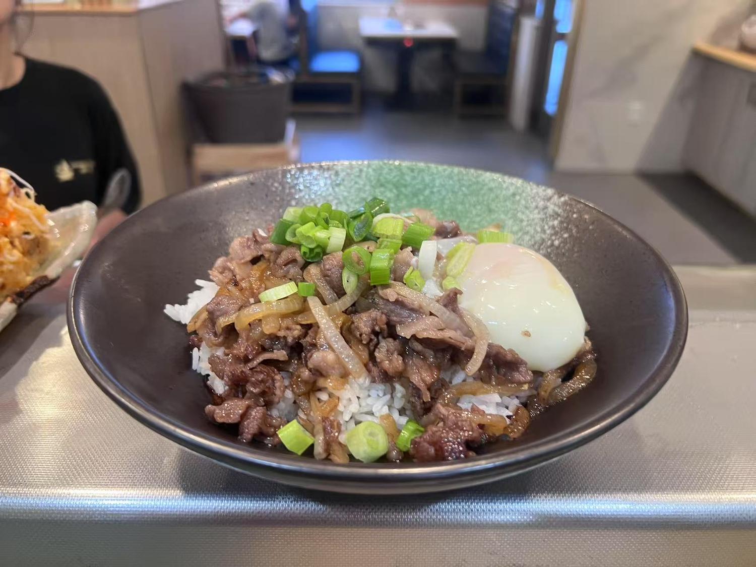 Rice bowl topped with tender beef, onions, scallions, and a soft - boiled egg.