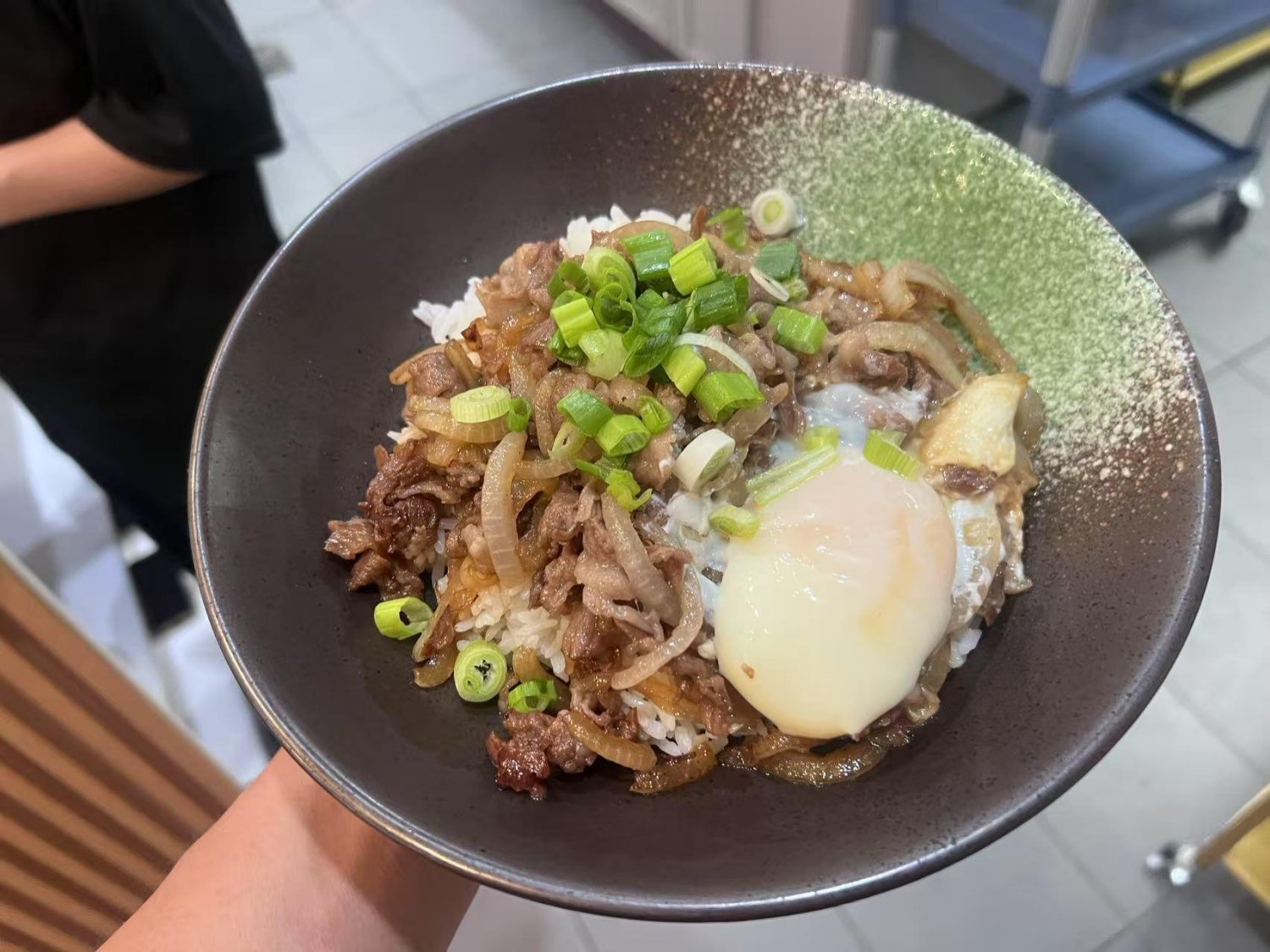 Hand - held rice bowl with seasoned beef, onions, scallions, and a runny egg on top.