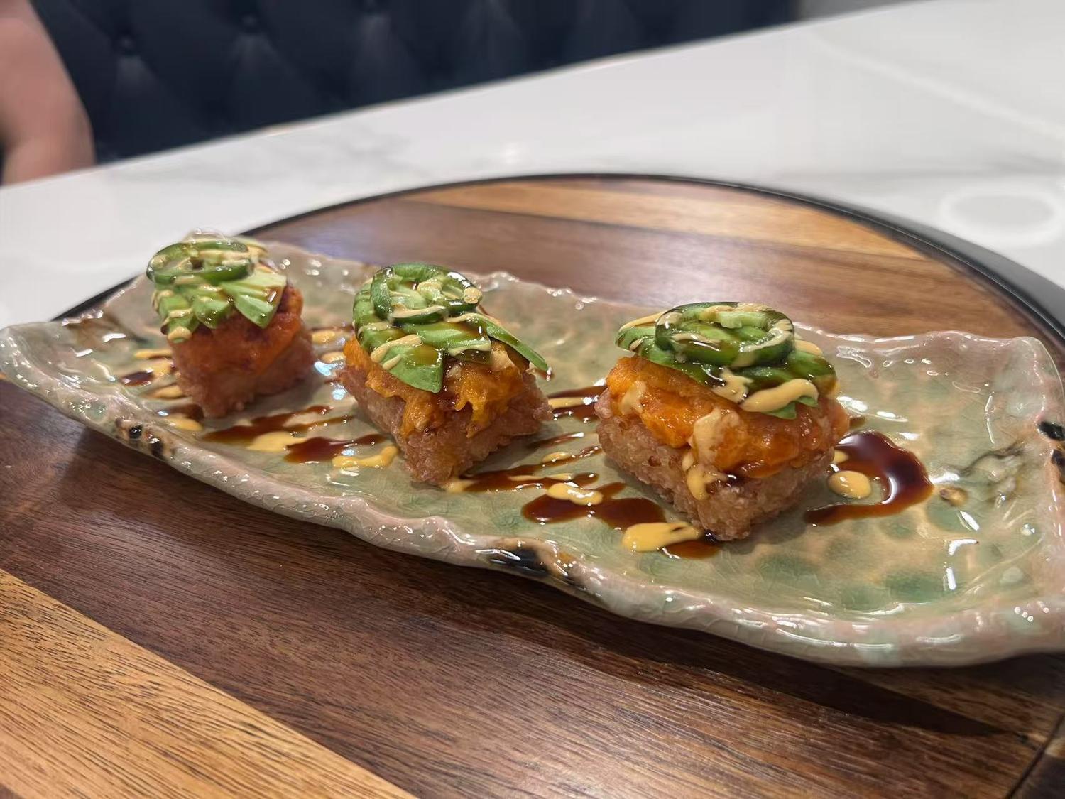 Three pieces of sushi topped with avocado, jalapeno, and sauce, arranged on a decorative plate.