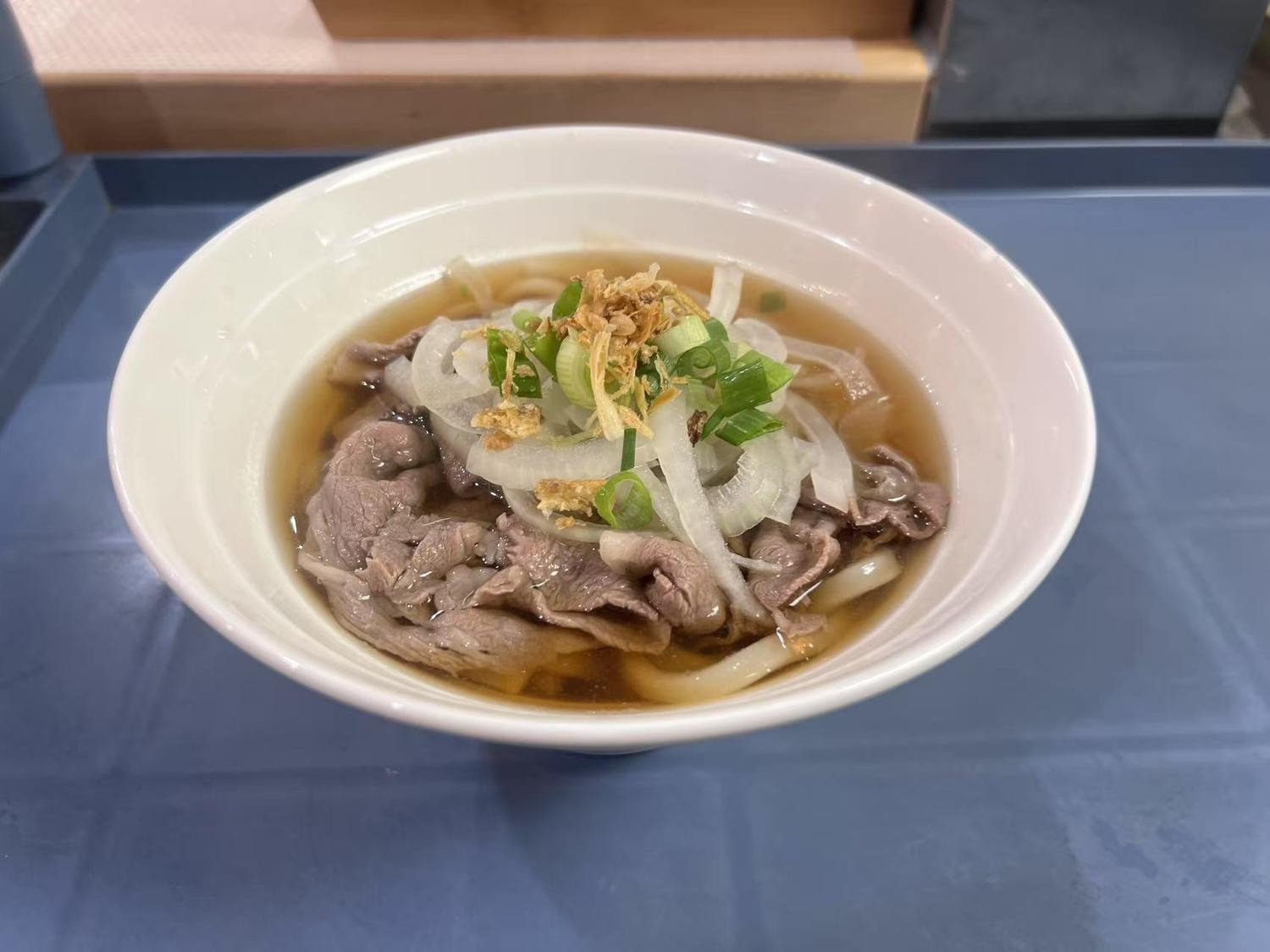 A bowl of udon soup with beef, onions, scallions and crispy bits, served on a blue tray.