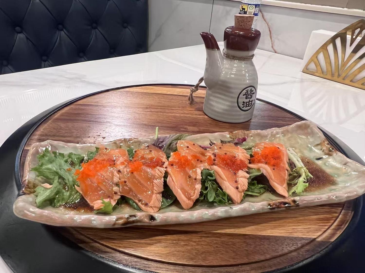 Salmon sashimi slices on lettuce with roe, paired with a soy sauce pitcher on a wooden board.