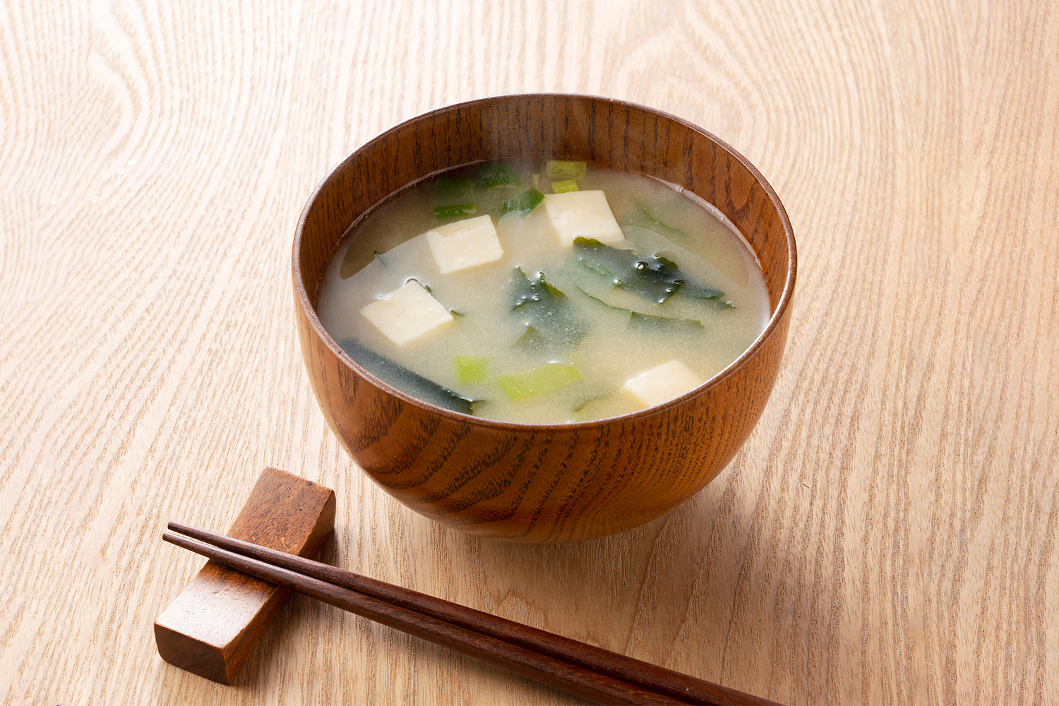 Wooden bowl of miso soup with tofu, seaweed, and chopsticks on a rest