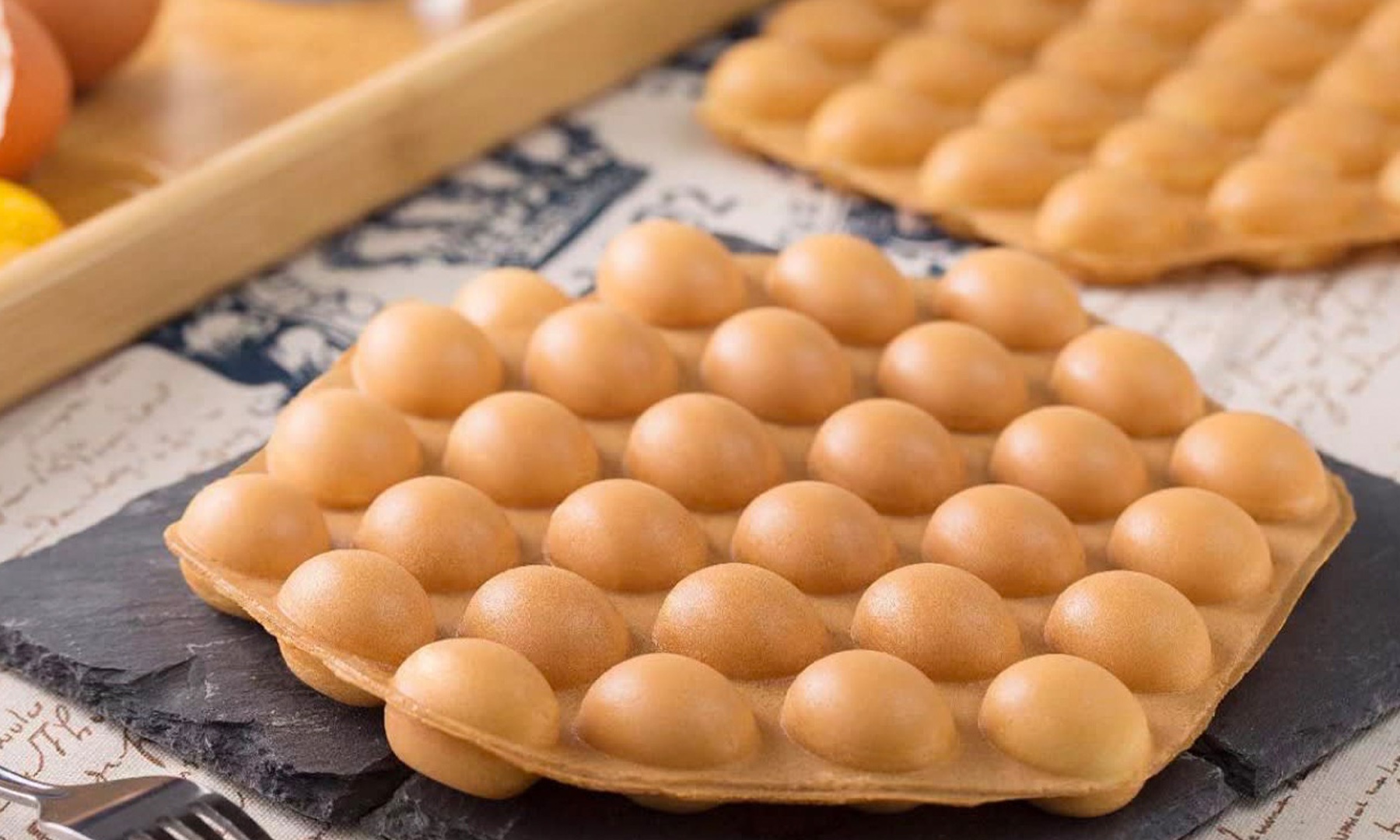 Plain golden‑brown bubble waffle rests on dark slate plate, classic round‑bubble texture, raw eggs in wooden tray in soft‑focus background.