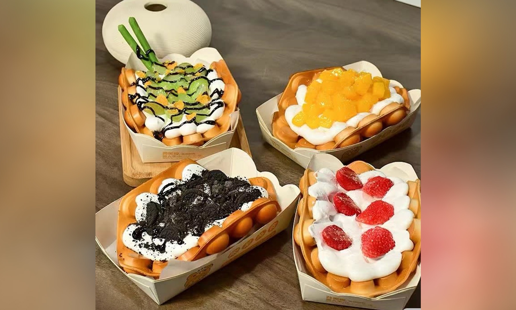 Four assorted bubble waffles in paper take‑out boxes, topped with whipped cream, fresh fruits, Oreo crumbs and chocolate drizzle on dark wooden table.