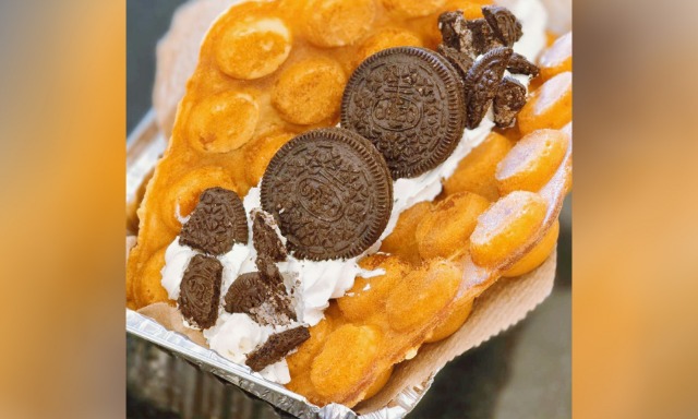 Close‑up of bubble waffle piled with whipped cream, whole and broken Oreo chocolate cookies, served in foil take‑out container.