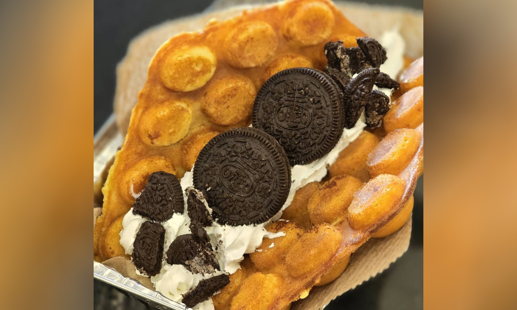 Golden bubble waffle topped with fluffy whipped cream, whole and crushed Oreo cookies, presented inside foil food container.