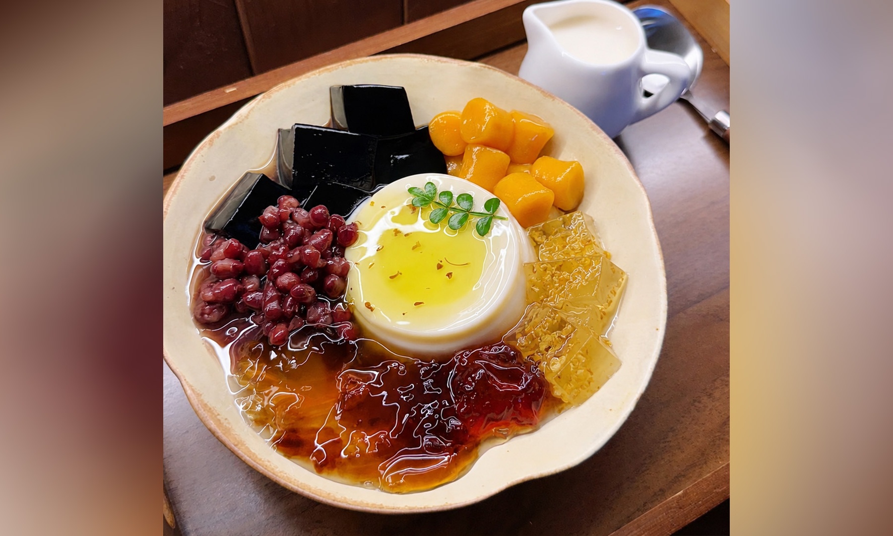 Colorful Chinese dessert bowl with milk pudding, grass jelly, red beans, taro balls, osmanthus jelly and sweet syrup, small milk pitcher on background.