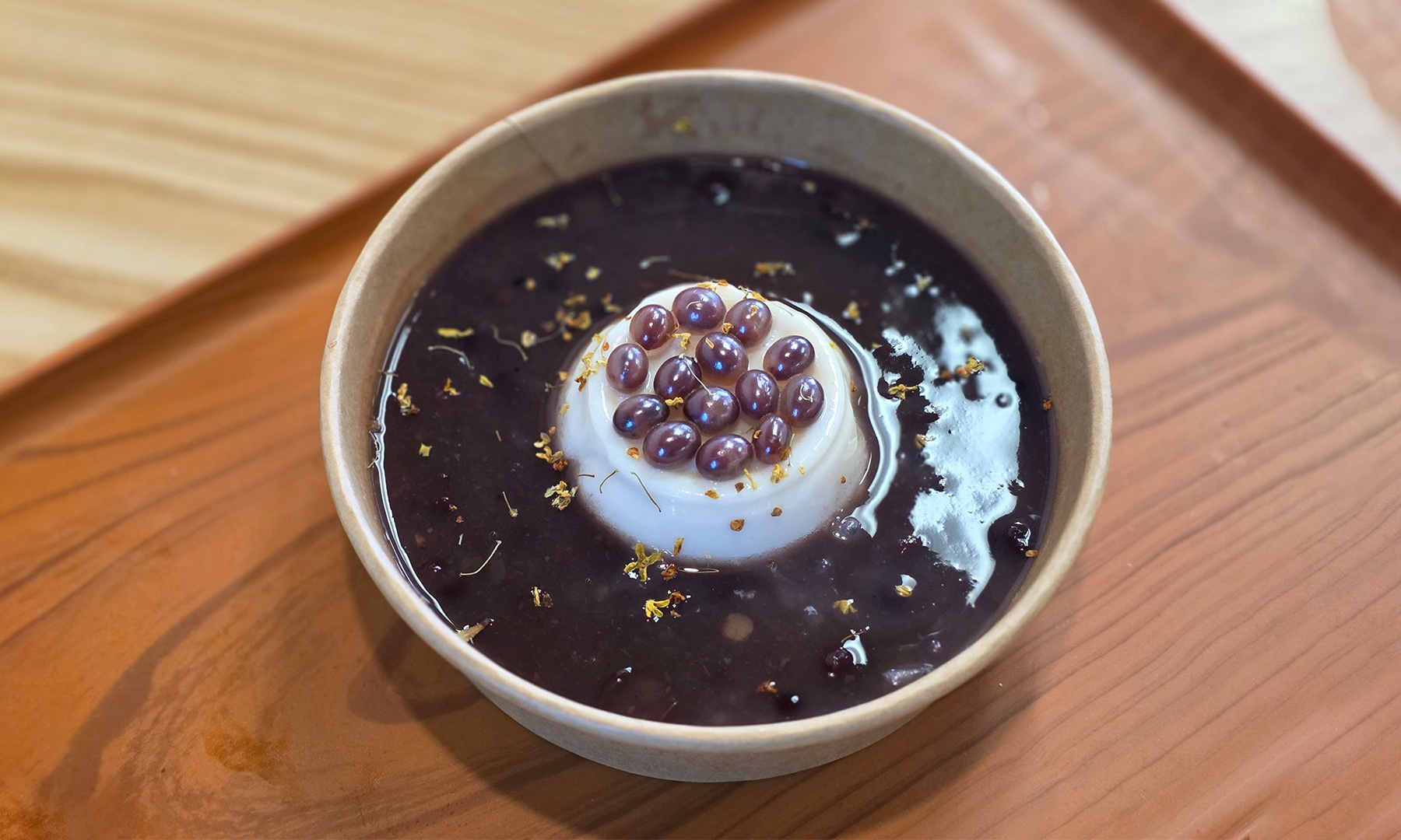 Bowl of thick red bean soup with white milk pudding center, popping boba on top, scattered osmanthus petals, placed on wooden tray.