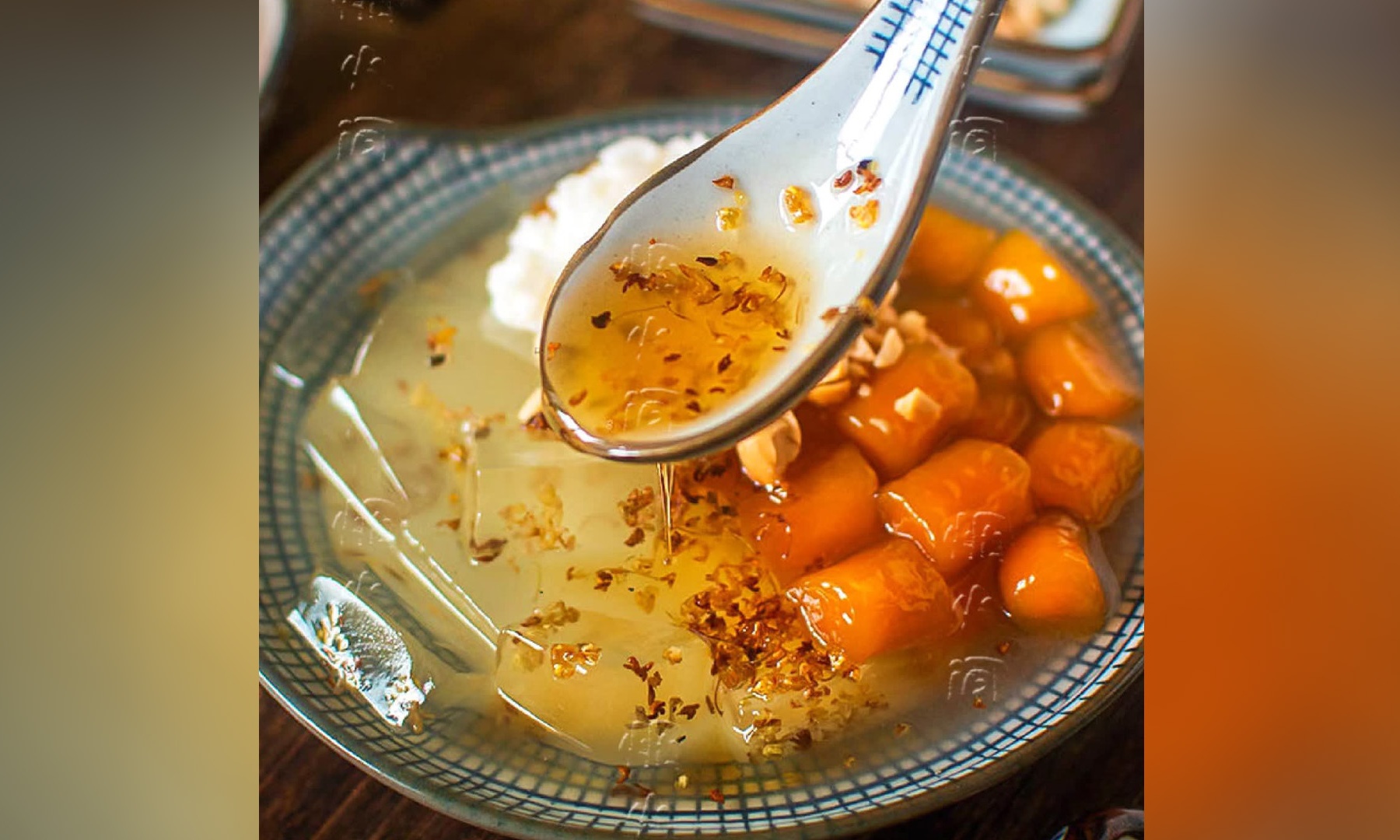Spoon pouring osmanthus honey syrup over crystal jelly and bright‑orange taro balls in blue‑grid ceramic bowl.