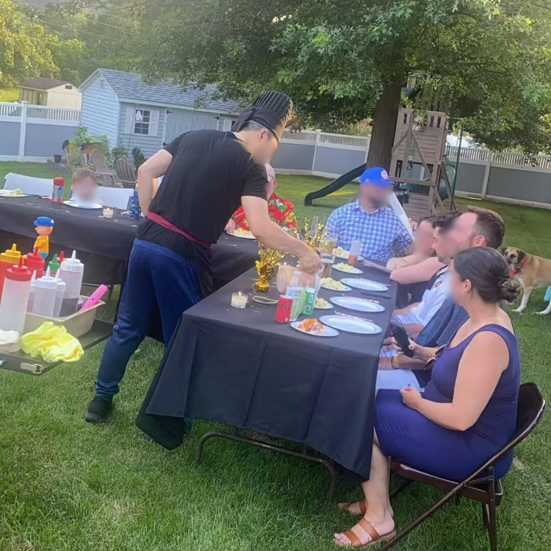 Hibachi chef serving food to guests at outdoor black - table setup.