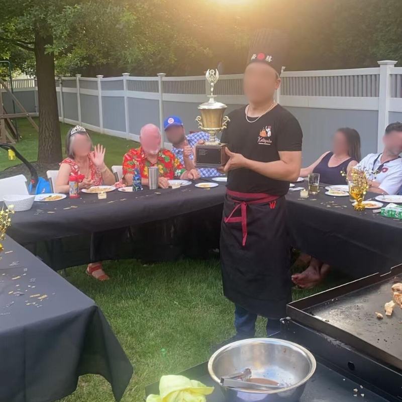 Hibachi chef holding trophy at outdoor party, guests seated at tables.