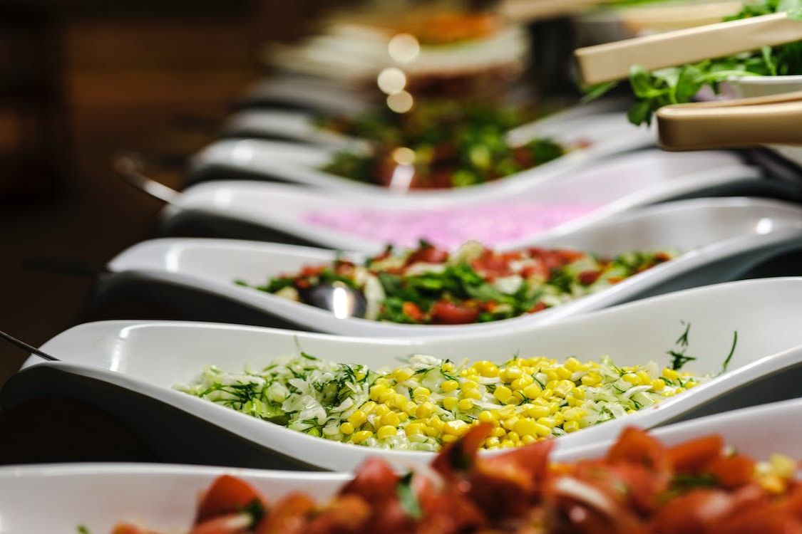 Row of buffet dishes with corn, tomatoes, and mixed vegetables.