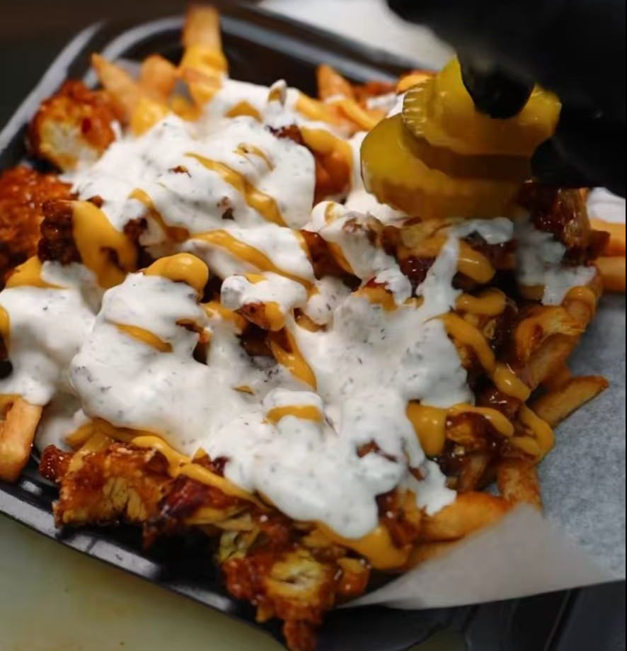 A close-up of loaded fries in a black takeout container, topped with crispy fried chicken, melted cheese sauce, and white ranch dressing. A gloved hand is adding pickles to the dish.