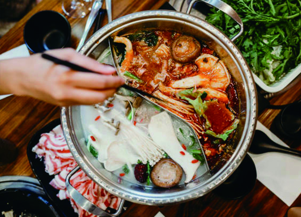 Hot pot with spicy and mild broth, featuring seafood, mushrooms, and vegetables, being enjoyed with chopsticks.