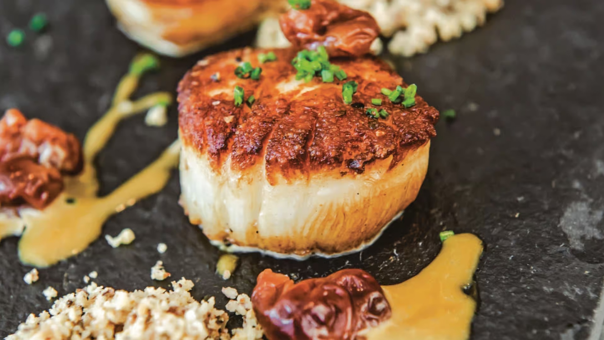 Seared scallop with bacon bits and sauce on a black plate