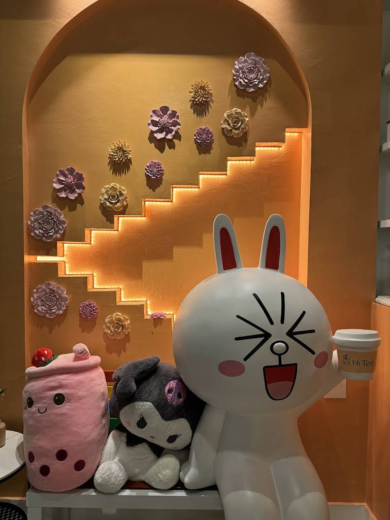 Hi-Tea-Bubble-Tea-Store-Interior-View-Of-The-Restaurant-Cape-Coral A cheerful white bunny statue holding a "Hi Tea" cup, sitting beside plush toys against a warm yellow wall with 3D flowers and glowing steps.
