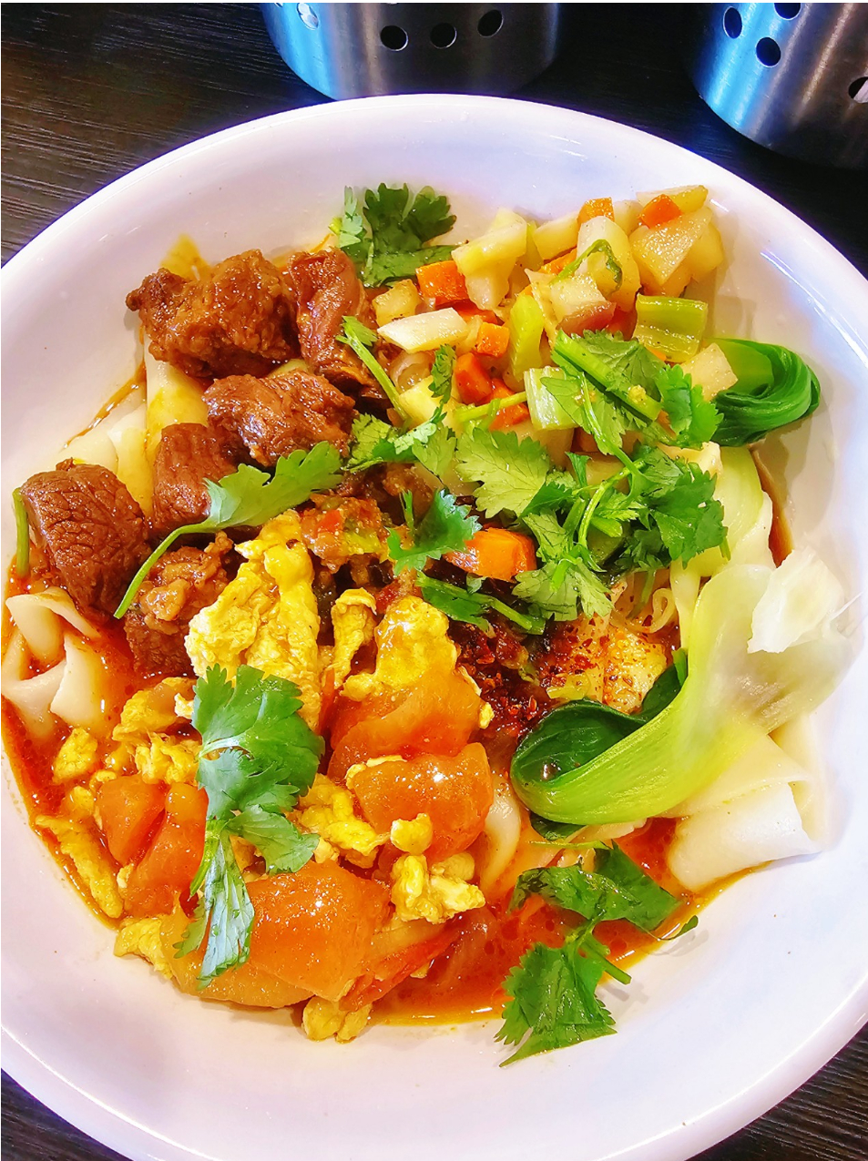 Bowl of wide noodles topped with beef, tomato egg, potatoes, and greens