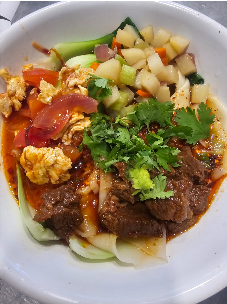 Bowl of wide noodles with beef, tomato egg, greens, potatoes, and cilantro