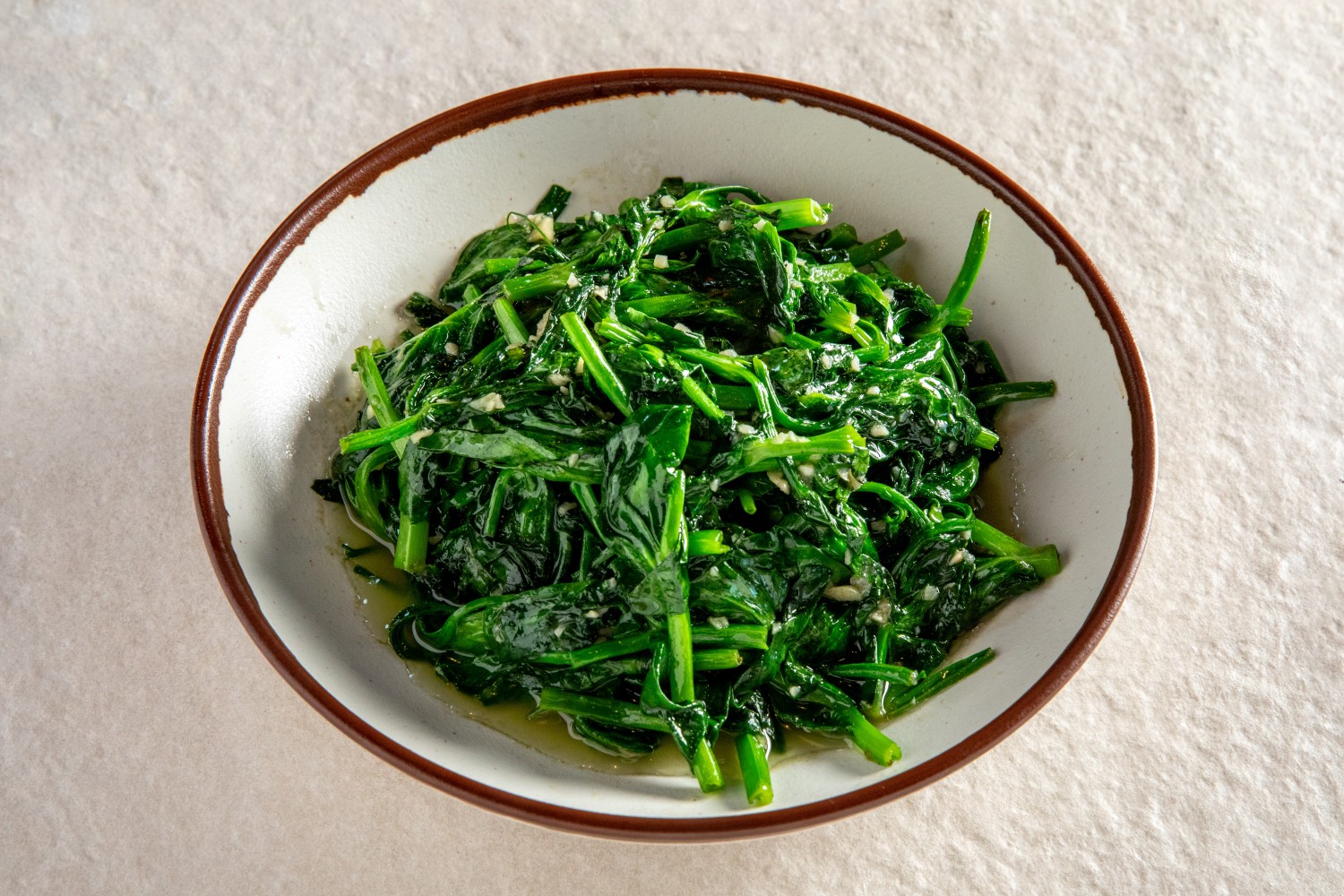 A bowl of garlic stir-fried leafy greens glistening with oil and minced garlic at Hunan Heat, a Chinese Restaurant in Long Island City