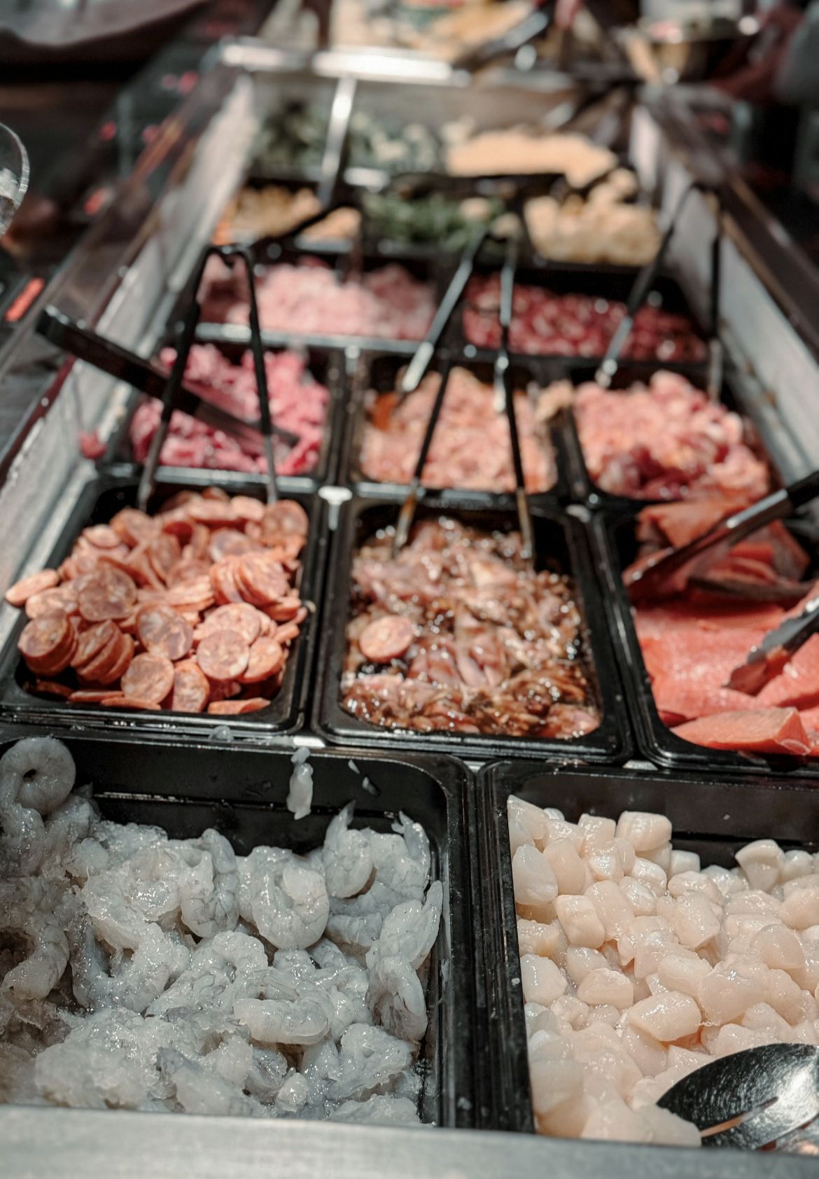 Close-up of a Mongolian grill ingredient bar with raw shrimp, scallops, sliced beef, sausage, and marinated meats.