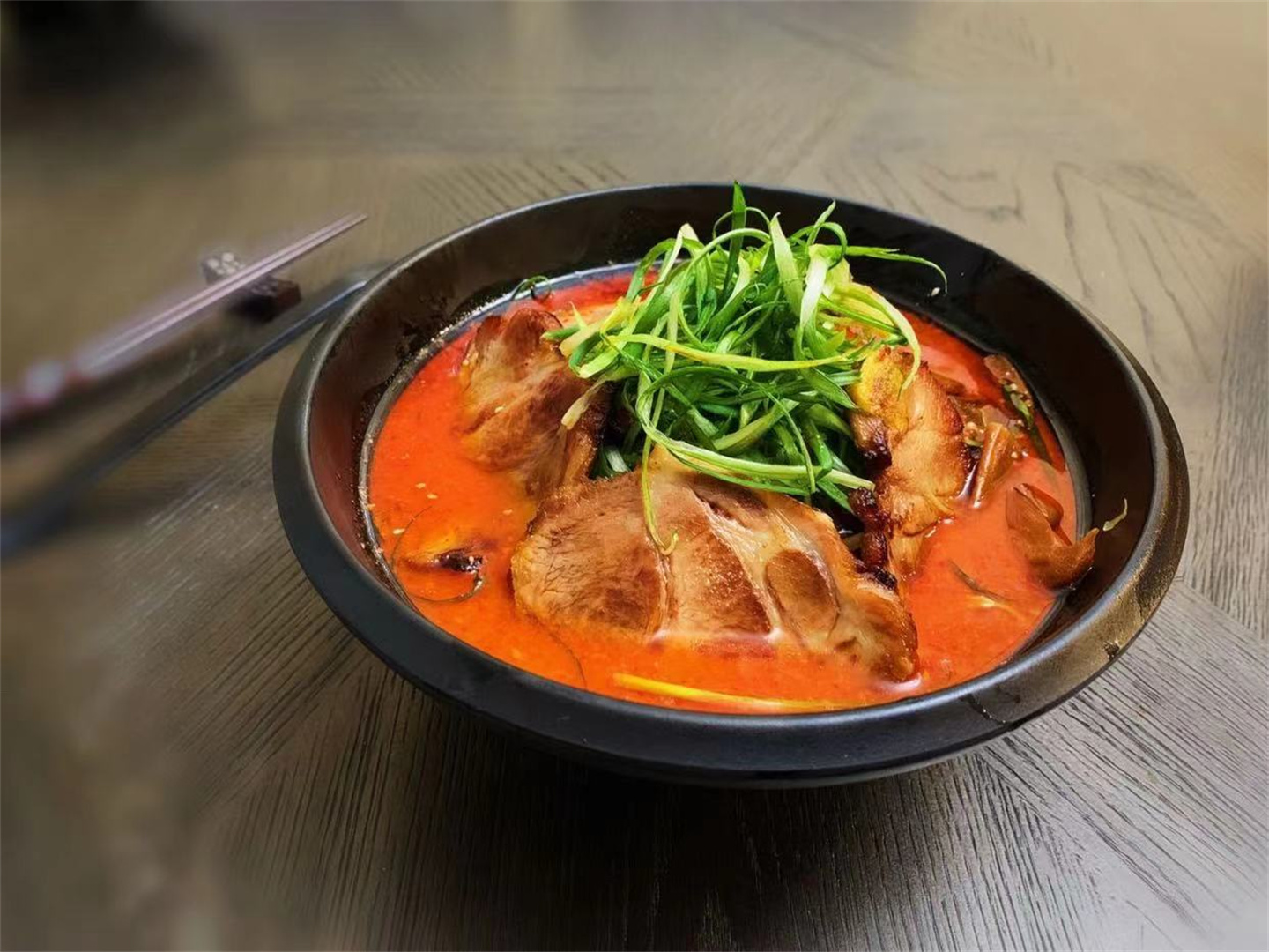 Spicy red‑broth ramen bowl topped with pork slices and fresh green scallions at Ichitaru Ramen, a Ramen restaurant in Carlsbad