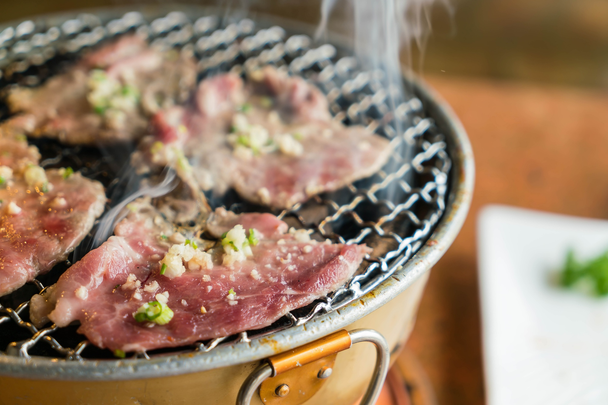 Thin raw beef slices seasoned with green onions grilling on metal mesh BBQ grill with rising steam at Jeju Island Korean BBQ and Sushi, a Korean Barbecue Restaurant in Pearland