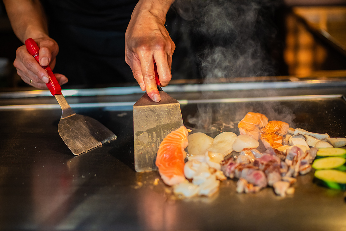 chef's hands with spatula over teppanyaki. cooking vegetables meat and seafood on hot hibachi grill table
