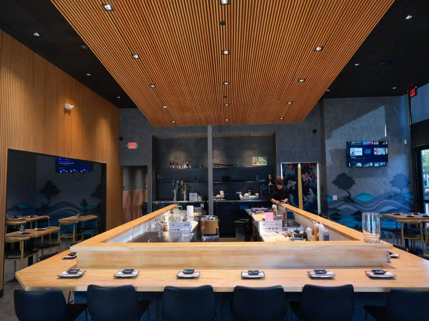 Modern sushi restaurant interior with a large wooden counter, slat ceiling, and artistic wall murals.