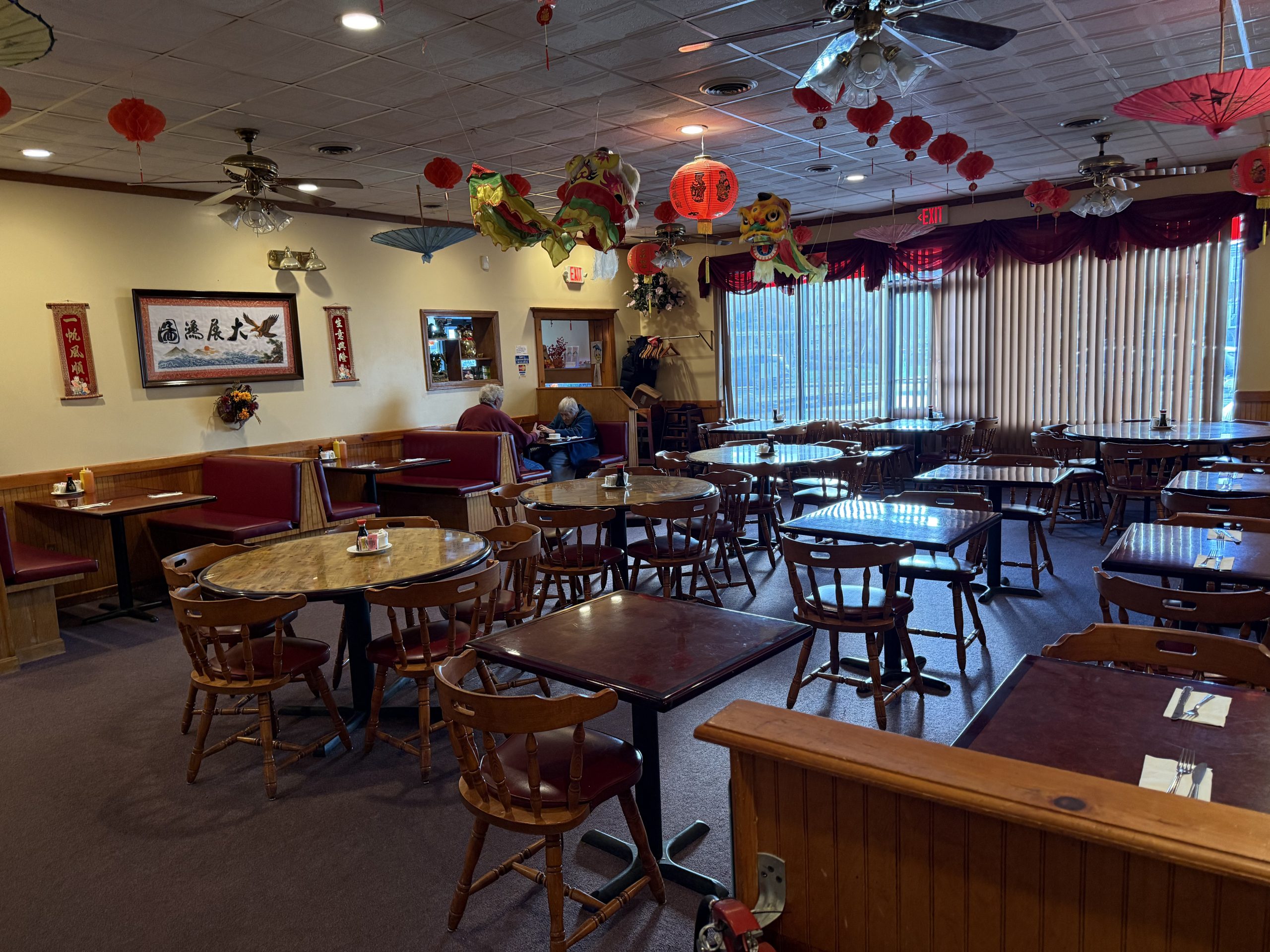 Kam’s-Cuisine-Chinese-Restaurant-Acushnet Interior of a Chinese restaurant with red lanterns and dragon dance decorations, wooden tables and chairs neatly arranged, calligraphy artworks on the walls, and two customers dining.