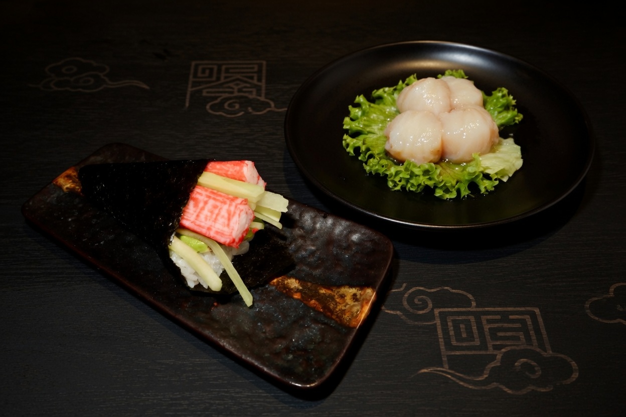 Sushi hand roll with crab and cucumber next to a small plate of raw scallops.