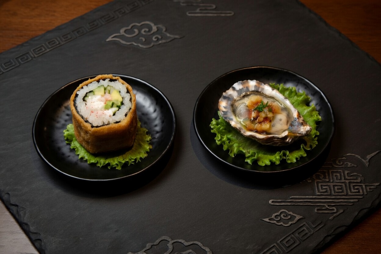 A tempura sushi roll and a dressed oyster on the half shell served on small black plates over a dark slate tray.