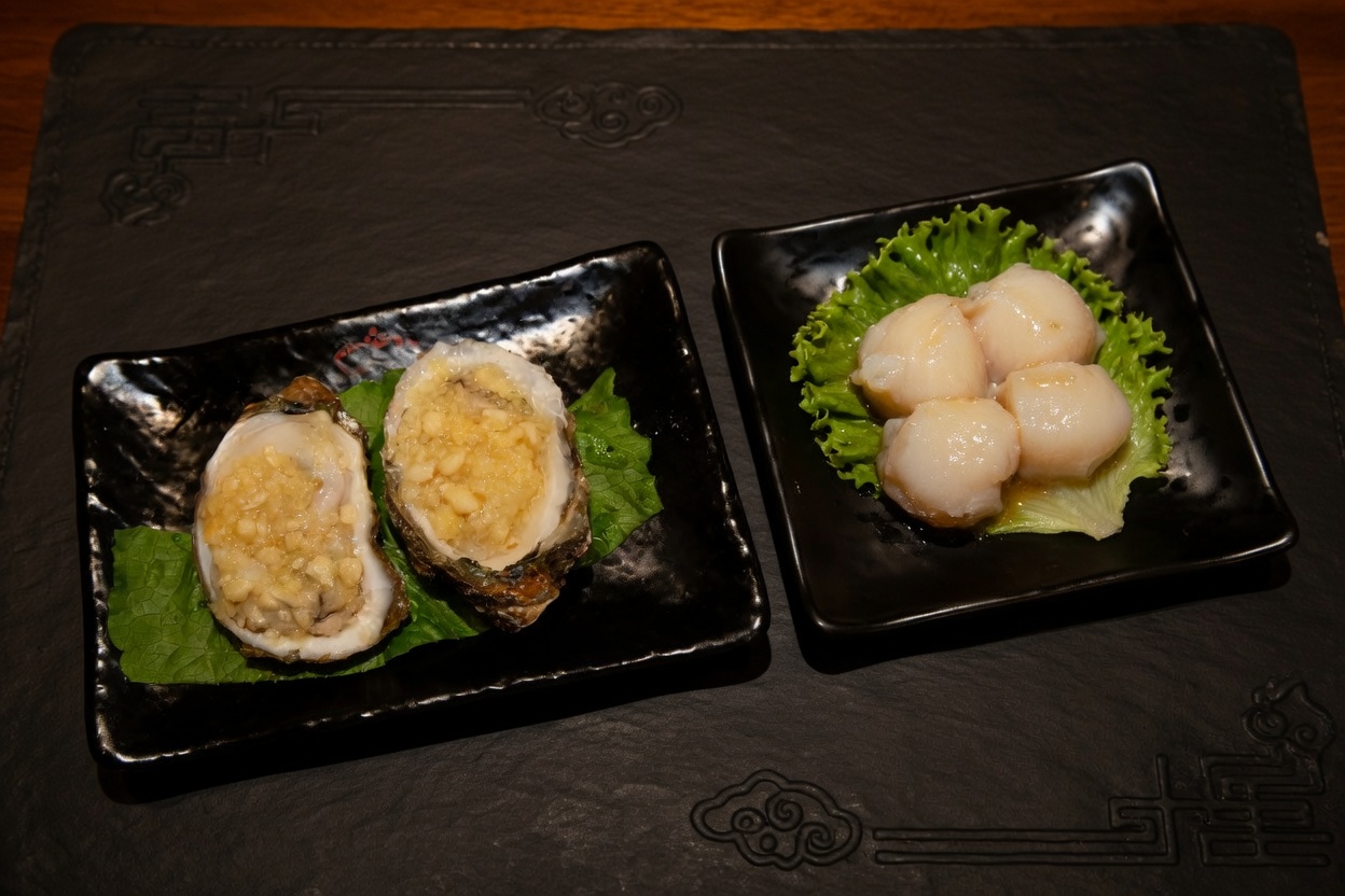 Two baked oysters with garlic topping and a serving of raw scallops on black plates.