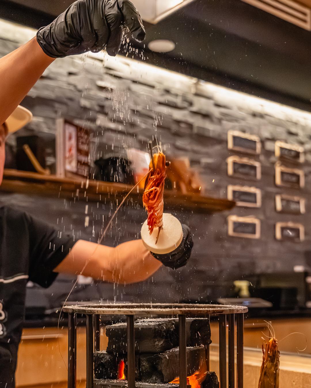 Chef seasons shrimp above burning charcoal grill at Ki Omakase, a Sushi Restaurant in Little Neck
