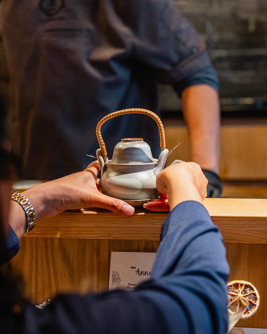 Guest gets woven-handle teapot at wooden counter at Ki Omakase, a Sushi Restaurant in Little Neck