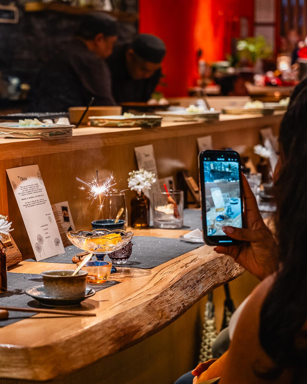Guest takes photo of sparkler-topped dessert at counter at Ki Omakase, a Sushi Restaurant in Little Neck