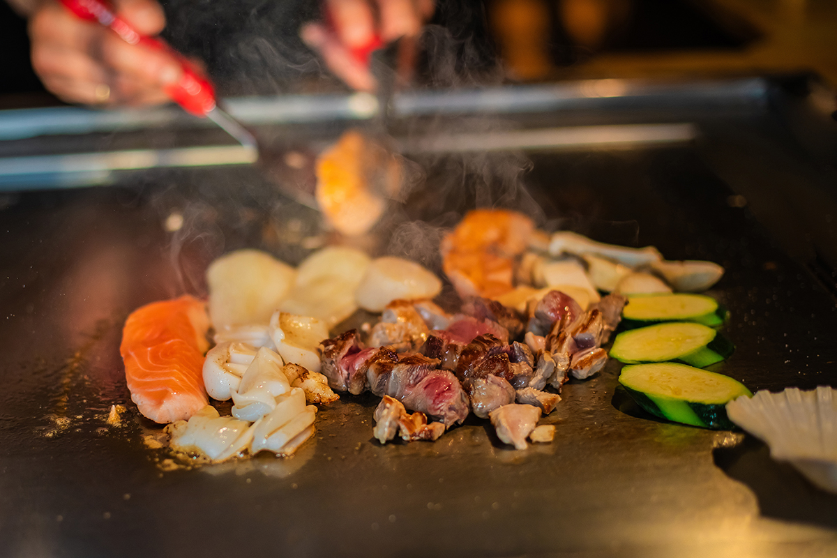 Kobe-Japanese-Steakhouse-Japanese-Restaurant-vegetables-meat-and-seafood-San-Antonio chef's hands with spatula over teppanyaki. cooking vegetables meat and seafood on hot hibachi grill table. Traditional Japanese Cuisine. Teppan show