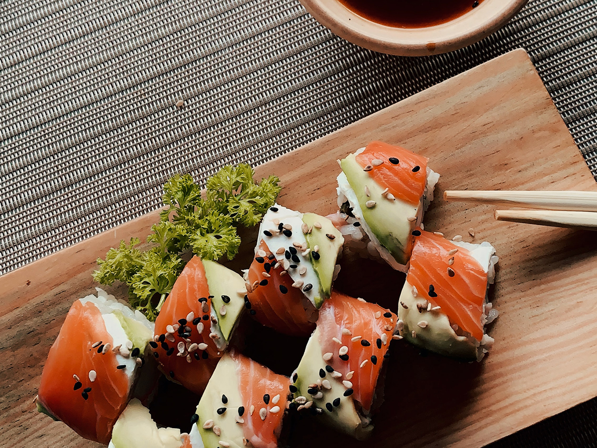 Top‑down shot of salmon avocado sushi rolls sprinkled with black and white sesame seeds on wooden board, garnished with parsley, chopsticks and small soy‑sauce bowl on woven mat.