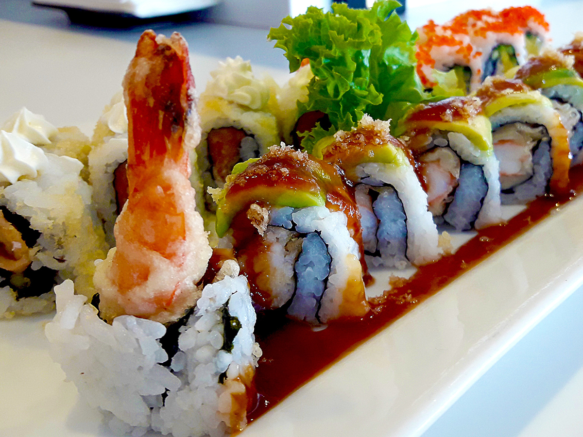 Variety of tempura shrimp sushi rolls on glossy white plate, topped with avocado and rich brown eel sauce, garnished with lettuce, drizzled sauce runs along edge of dish.
