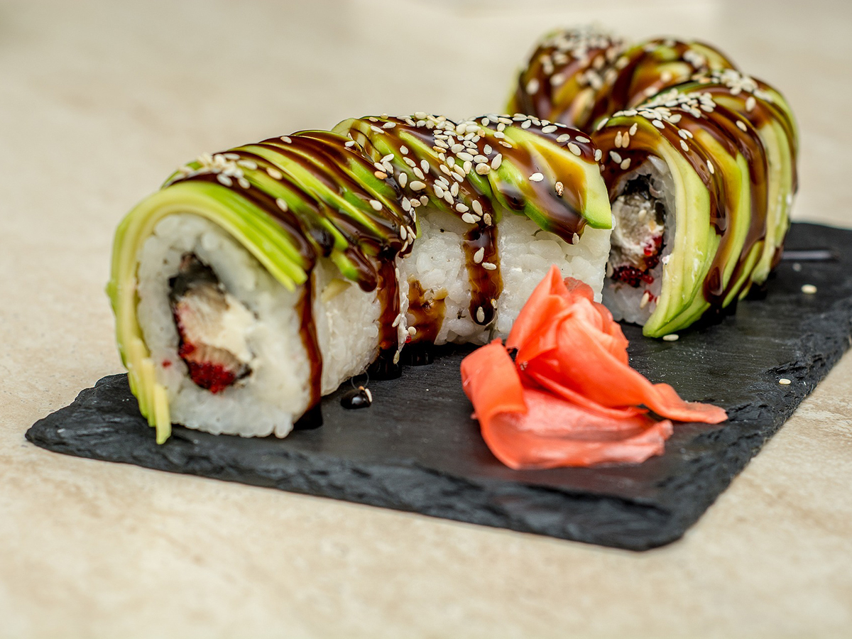 Avocado‑covered sushi rolls glazed with dark brown sauce and sesame seeds, served on dark slate plate, pickled ginger placed beside the sliced sushi pieces.