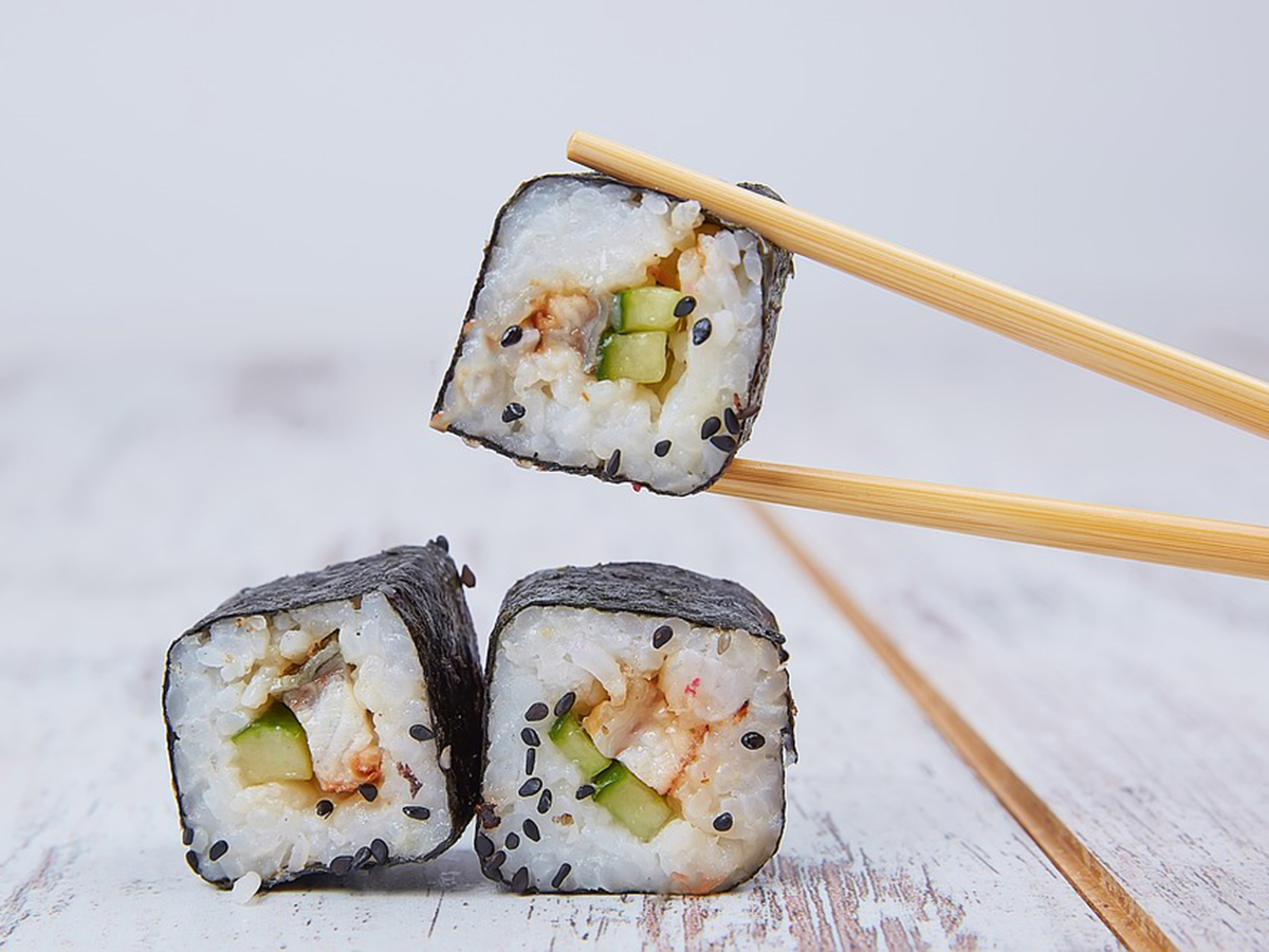 One piece of seaweed maki roll held by wooden chopsticks, two more pieces rest on weathered white wooden table, filled with cucumber and fish, sprinkled with black sesame seeds.