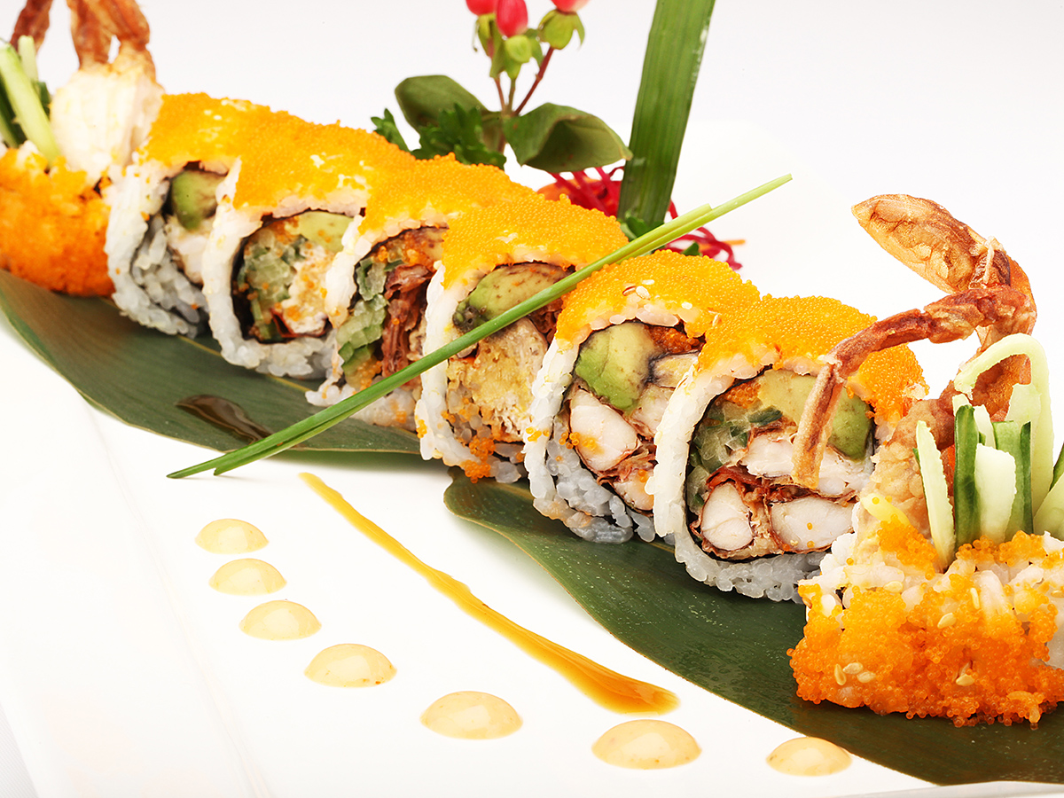 Sushi rolls covered with bright orange fish roe, soft‑shell crab pieces at ends, laid on green banana leaf, decorated with herbs and artistic sauce drizzles on white plate.